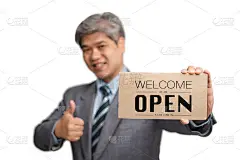 亚洲商人不戴医用口罩就在孤立的背景下，举着欢迎我们的牌子。经营理念和店铺都要开业后情况才会恢复正常。