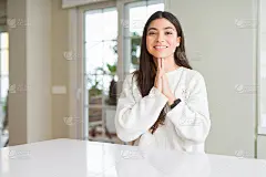 年轻美丽的女人在家里白色的桌子上双手合十祈祷，自信地微笑着请求原谅。