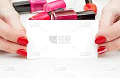 女性的手与红色美甲拿着名片和瓶子与指甲油在白色木制桌子上。