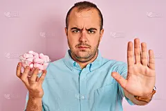 年轻男子拿着碗与棉花糖站在孤立的粉红色背景与开放的手做停止标志与严肃和自信的表情，防御手势