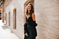 Pretty young girl with brunette wavy hair bright makeup offshoulder dress and black jacket holding glasses in hands walking at daylight city and smiling beige building background