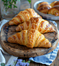 top view of three golden brown croissants on an old wooden board, blue fabric serving napkin in white cell / some on the side and one in front with gold accents, next to it is another platter full of other french pastries such as tarts or charcuterie boar