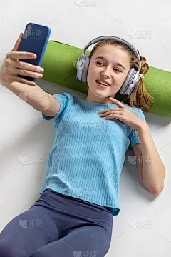 瑜伽结束后，女孩戴着耳机拿着手机休息。绿色的地毯滚在你的头下。