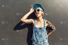 自由泳。另类女孩戴着帽子和太阳镜孤立地站在灰色的城市街道上，显示舌头开玩笑