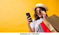 Shopaholic young Asian woman wearing hat and sunglasses with holding shopping bag, She smiling happy using smartphone for shopping in yellow background. 库存照片