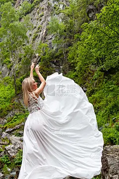 一个年轻的金发女郎摆着优雅的姿势，在群山中撩起一件闺房裙，靠在瀑布上，石头举起她的双手，就像一个温暖